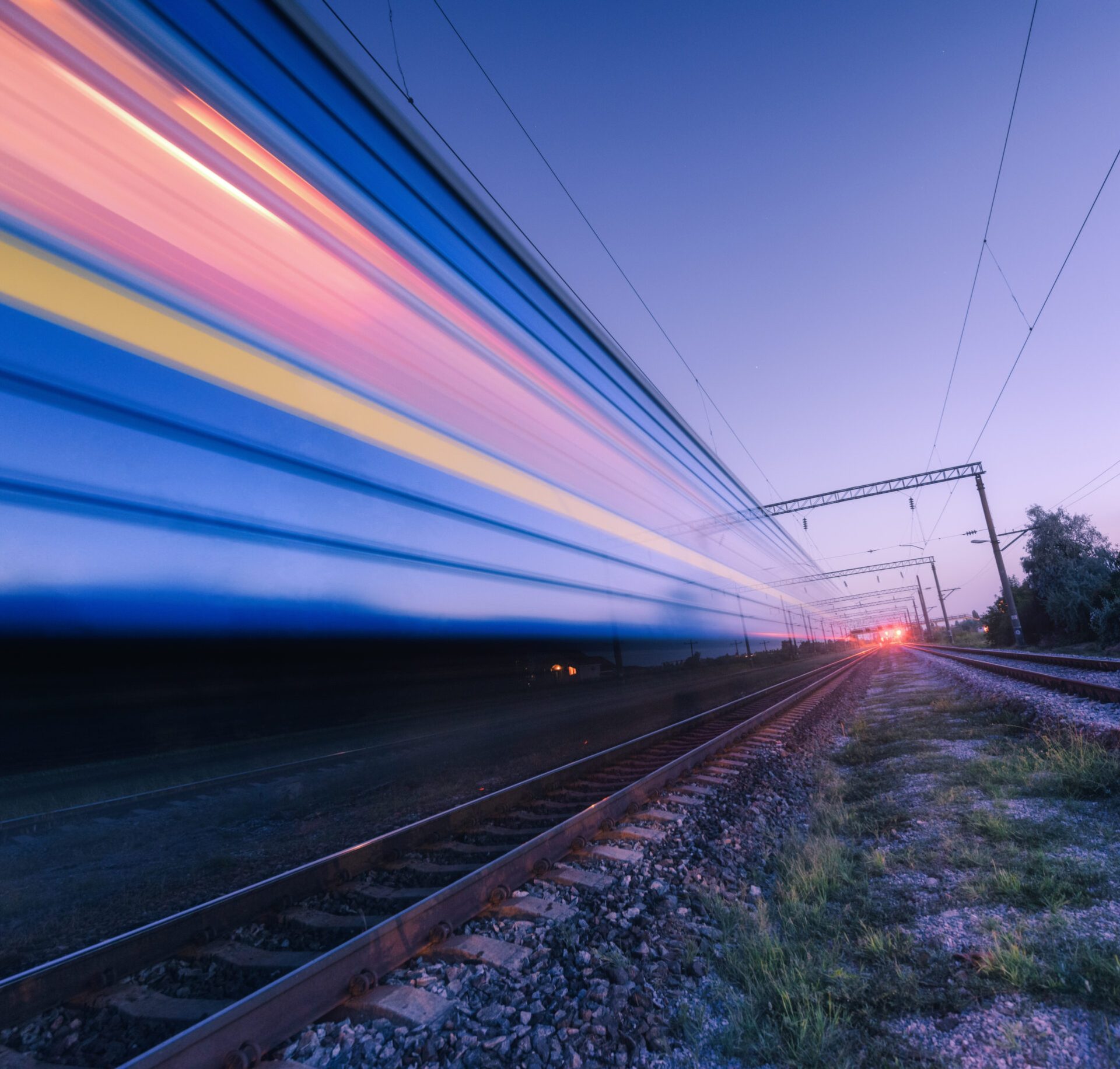 High speed passenger train in motion on the railroad at summer night. Moving blurred modern commuter train at dusk. Industrial landscape with railway station and purple sky. Transport.  Travel