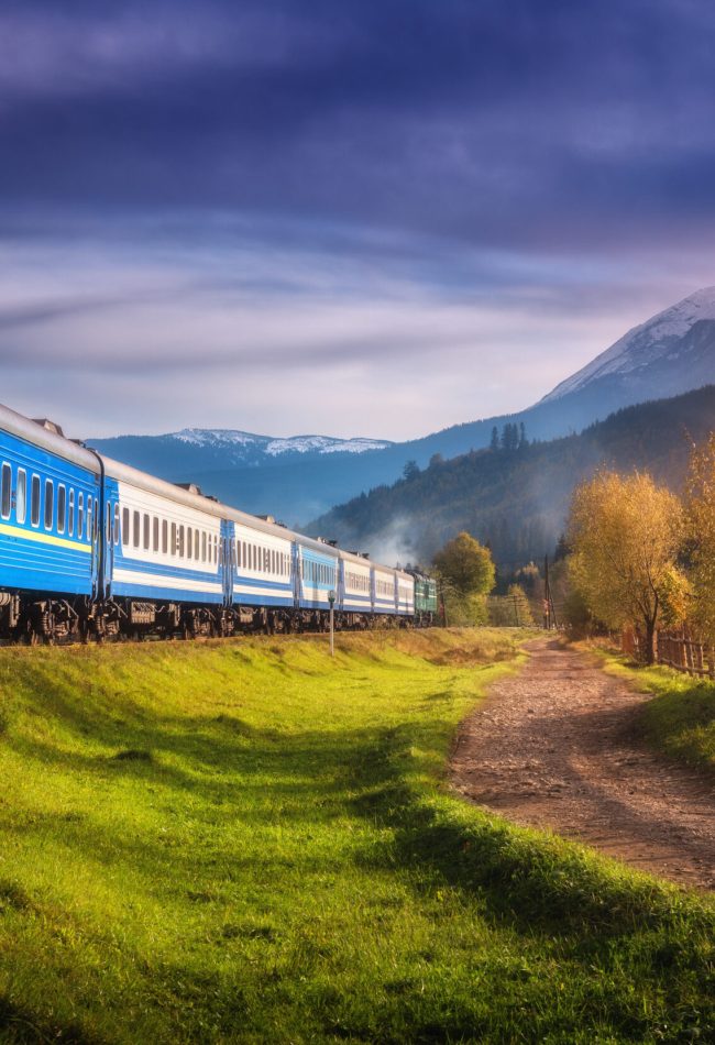 Moving train in mountains at sunset in autumn. Industrial landscape with passenger speed train on railroad, dirt road, snowy rocks, orange trees, green grass, purple sky in fall. Rural railway station